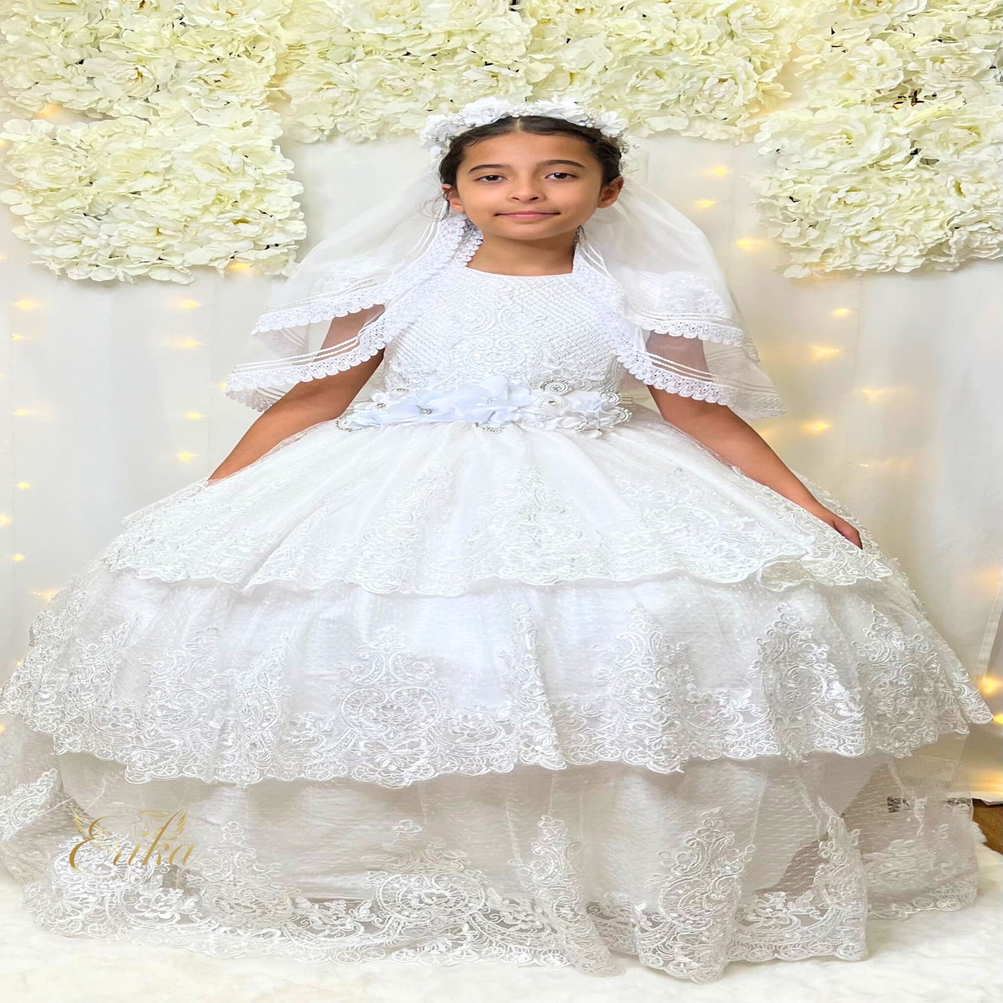 Front view of a white First Communion dress with three tiered lace layers and a colorful floral sash